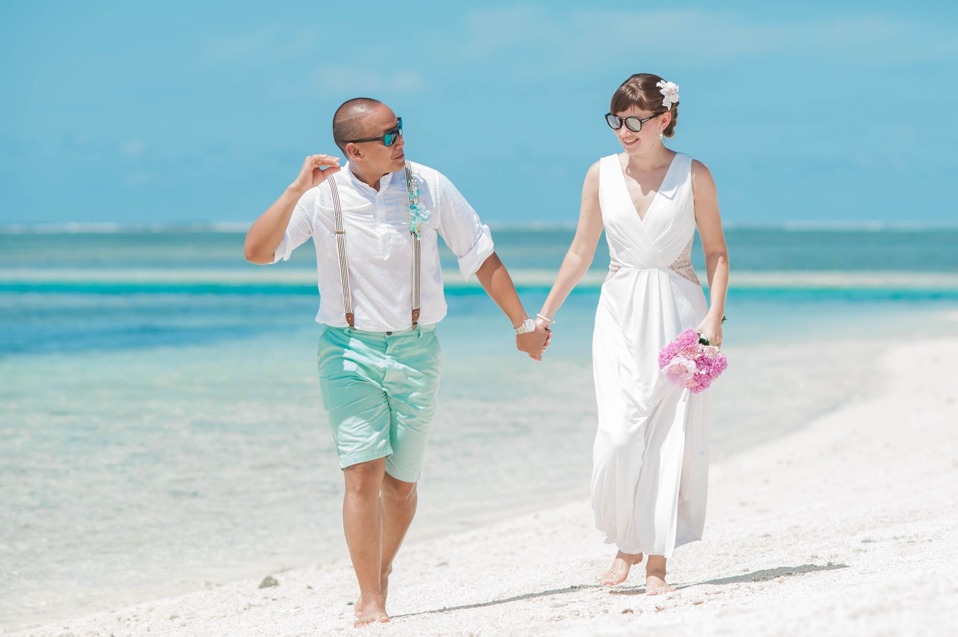 A joyful couple holding hands, walking on a sunny beach in wedding attire.