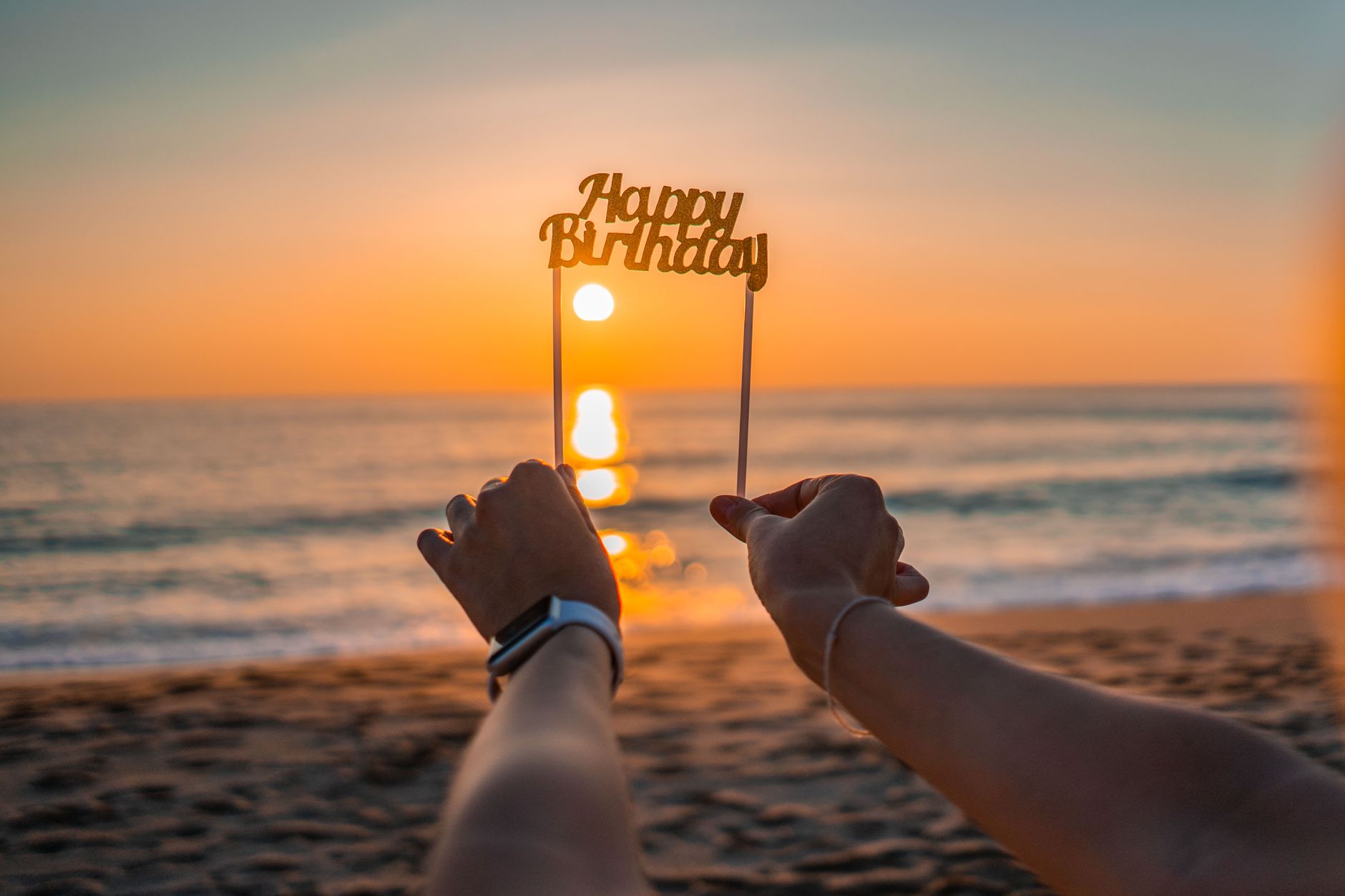 Hands holding 'Happy Birthday' sign against a vibrant beach sunset.