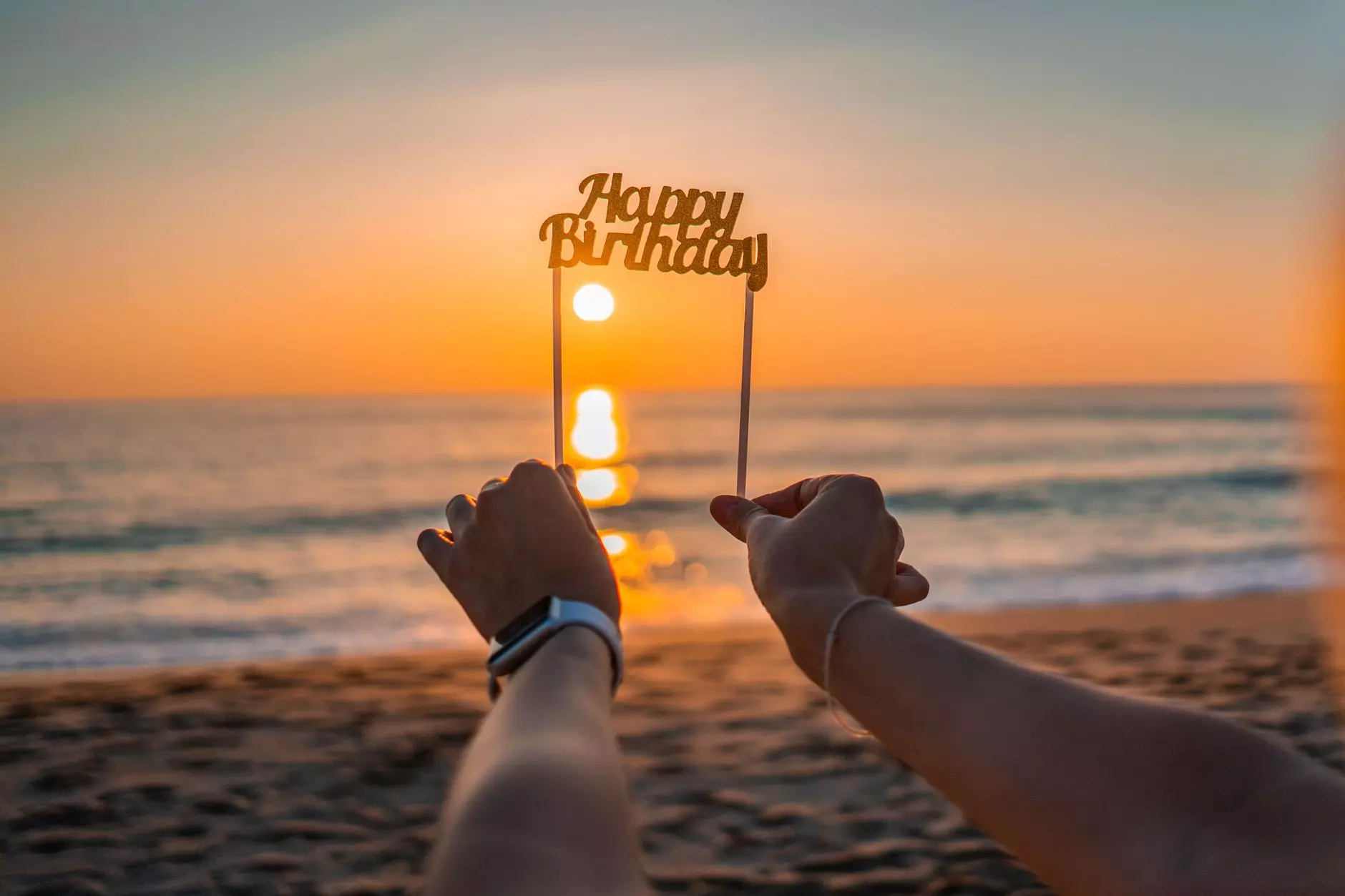 Hands holding 'Happy Birthday' sign against a vibrant beach sunset.