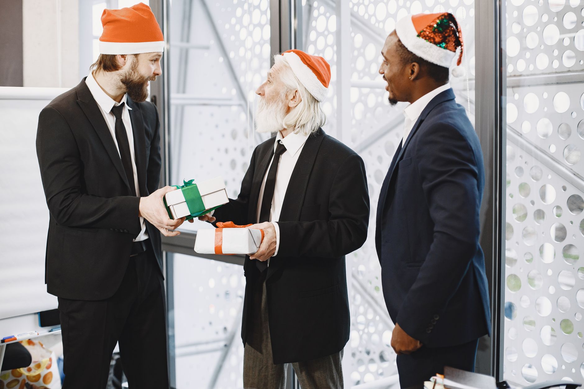 Three businessmen in Santa hats exchanging gifts in a festive office environment.
