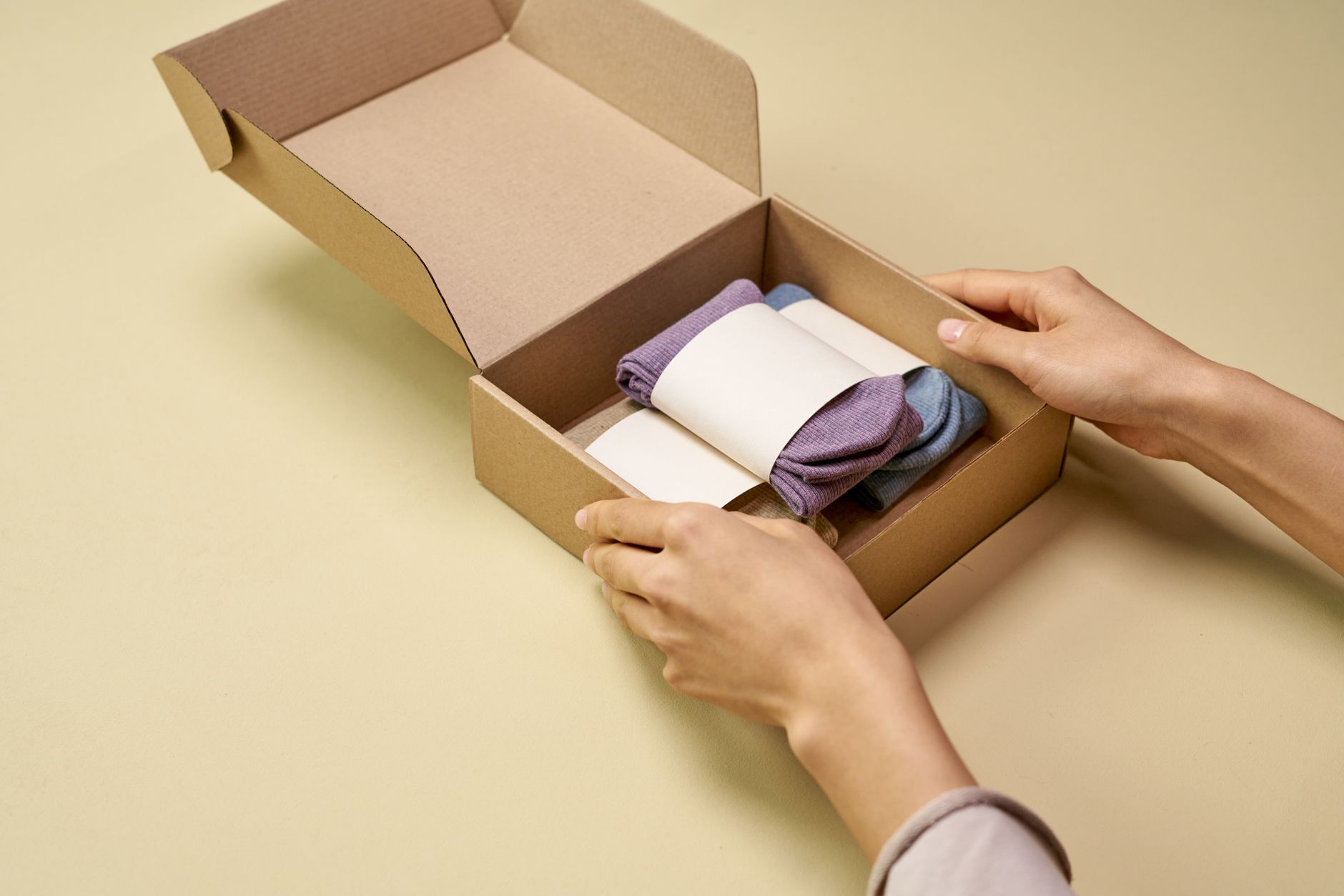 A person carefully places folded socks into a cardboard packaging box on a beige background.