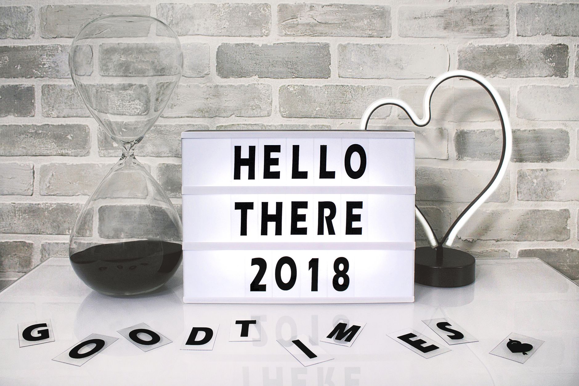 Contemporary design scene with a lightbox message, hourglass, and heart-shaped lamp on a brick wall background.