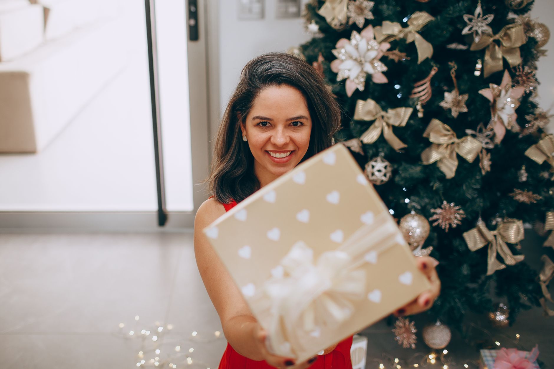 Smiling woman holding a Christmas gift in front of a decorated tree indoors.