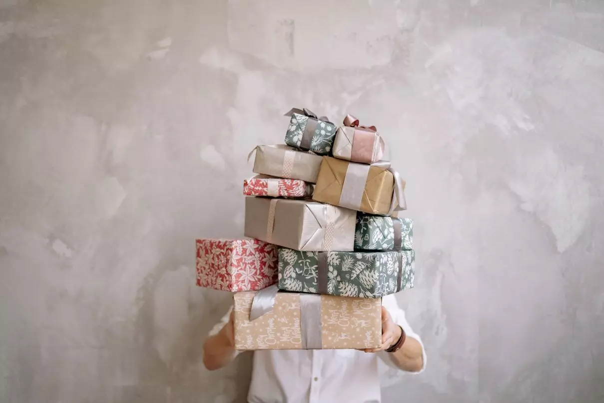 Person hiding behind a tall stack of gift-wrapped boxes against a gray wall, indoors.