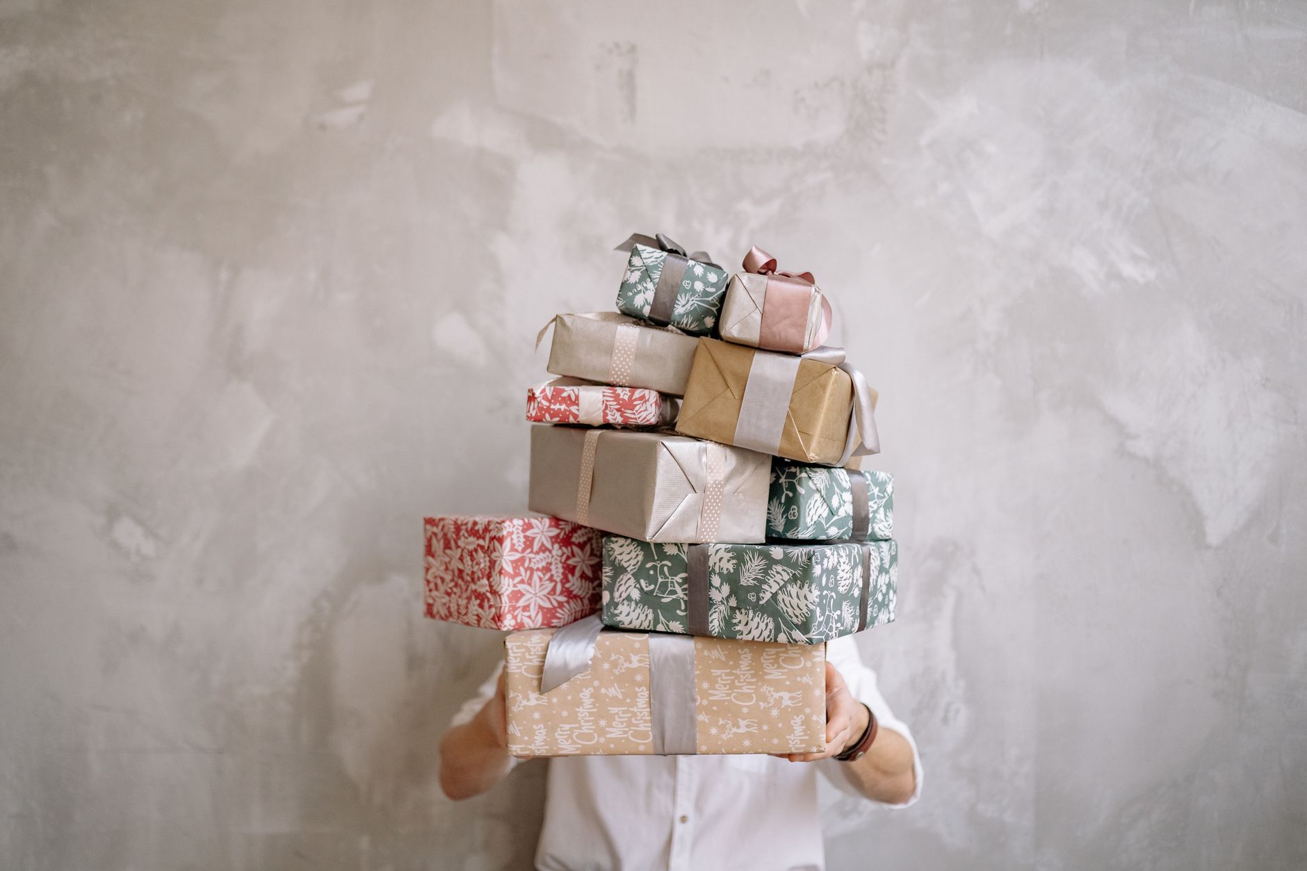 Person hiding behind a tall stack of gift-wrapped boxes against a gray wall, indoors.