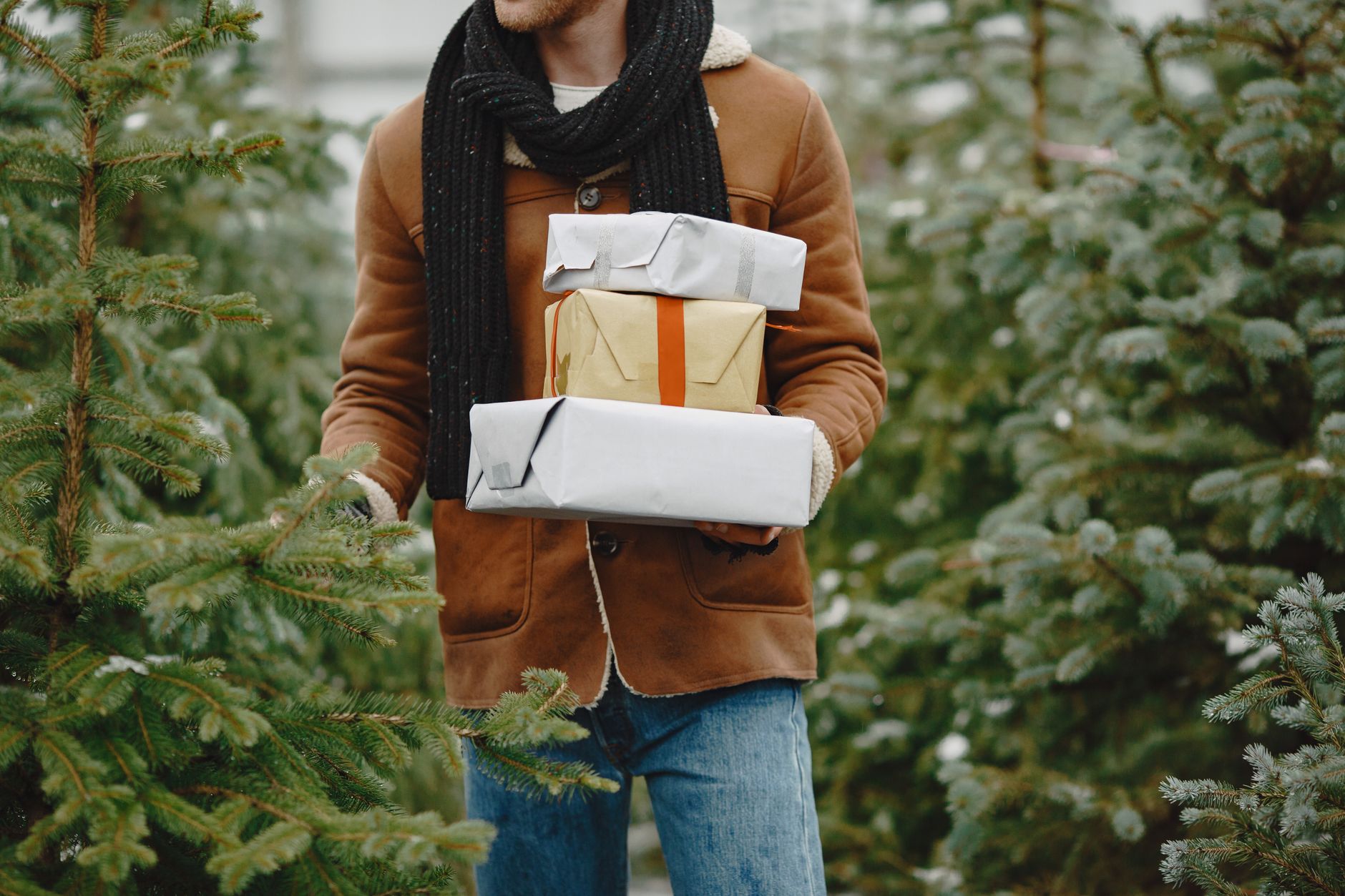 A person in a brown jacket holds gift boxes while standing in a forest of Christmas trees.