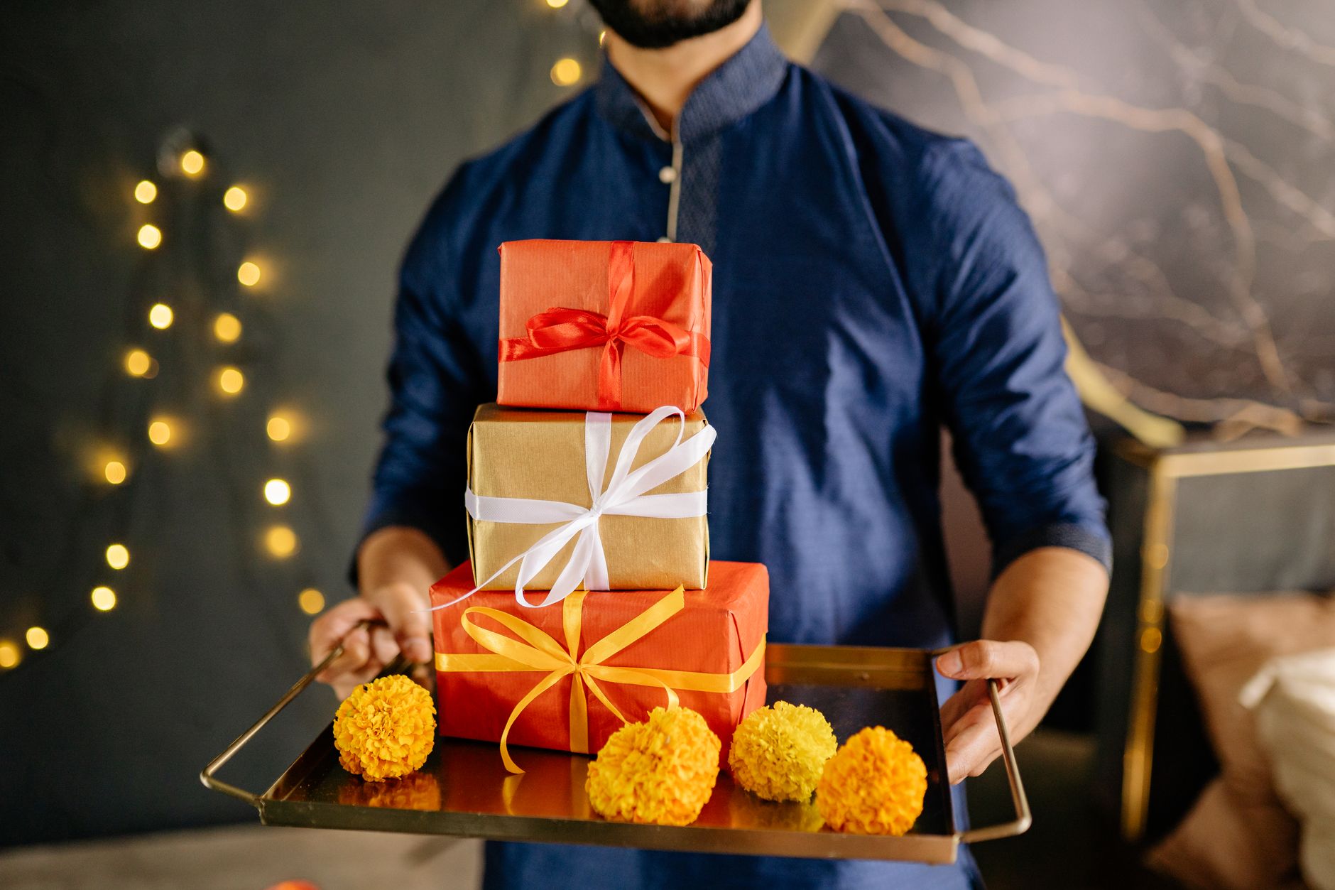 A man in traditional attire holding Diwali gifts with marigold flowers and festive lights.