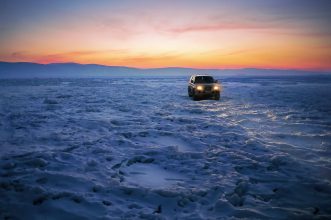 A vehicle drives on a frozen lake in Khuzhir, Russia at sunset with glowing headlights.