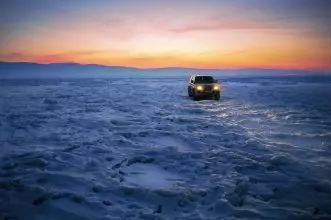 A vehicle drives on a frozen lake in Khuzhir, Russia at sunset with glowing headlights.