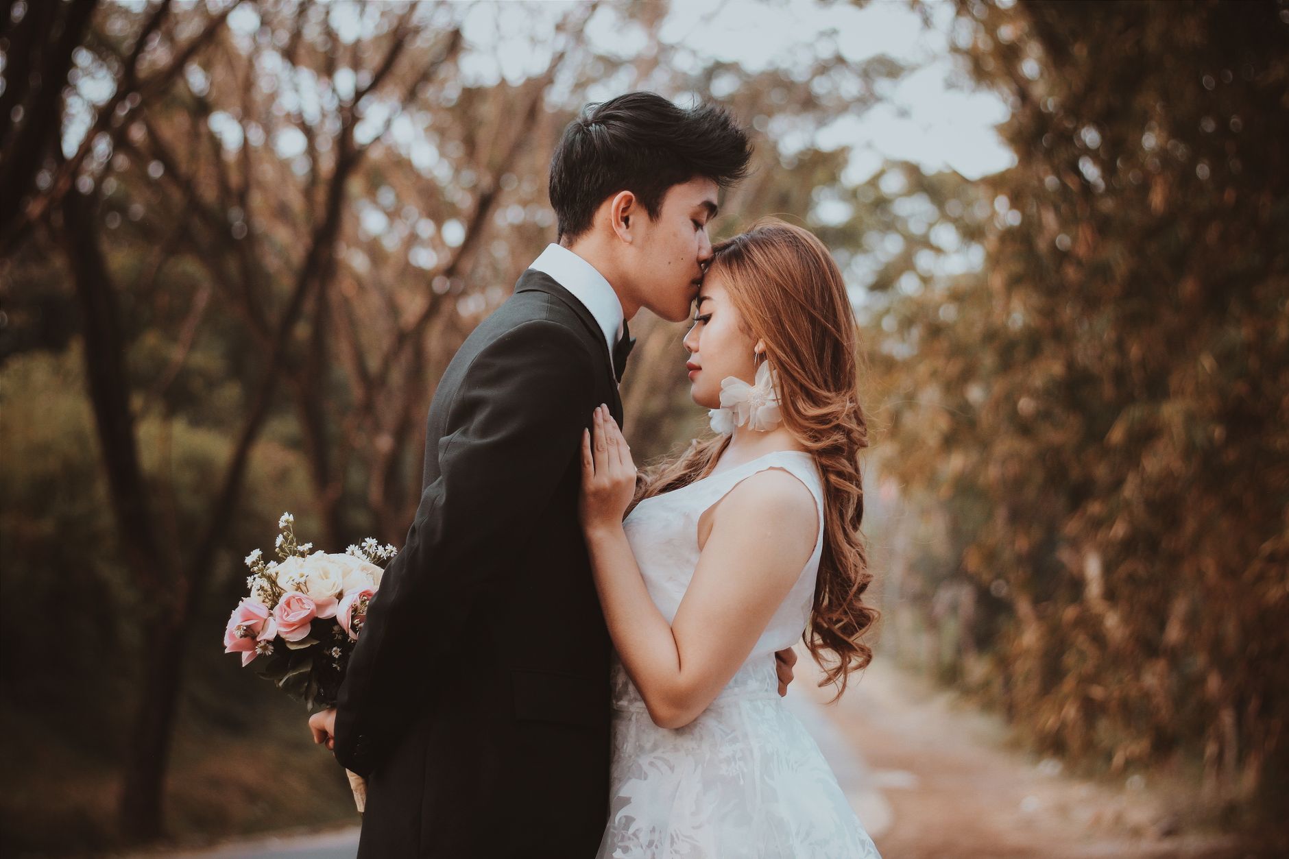 Side view of stylish ethnic groom in suit with bridal bouquet kissing gently charming bride in park