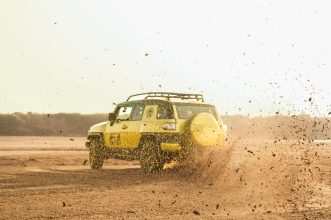Yellow SUV creating a dramatic display of dust while driving off-road in a desert setting.