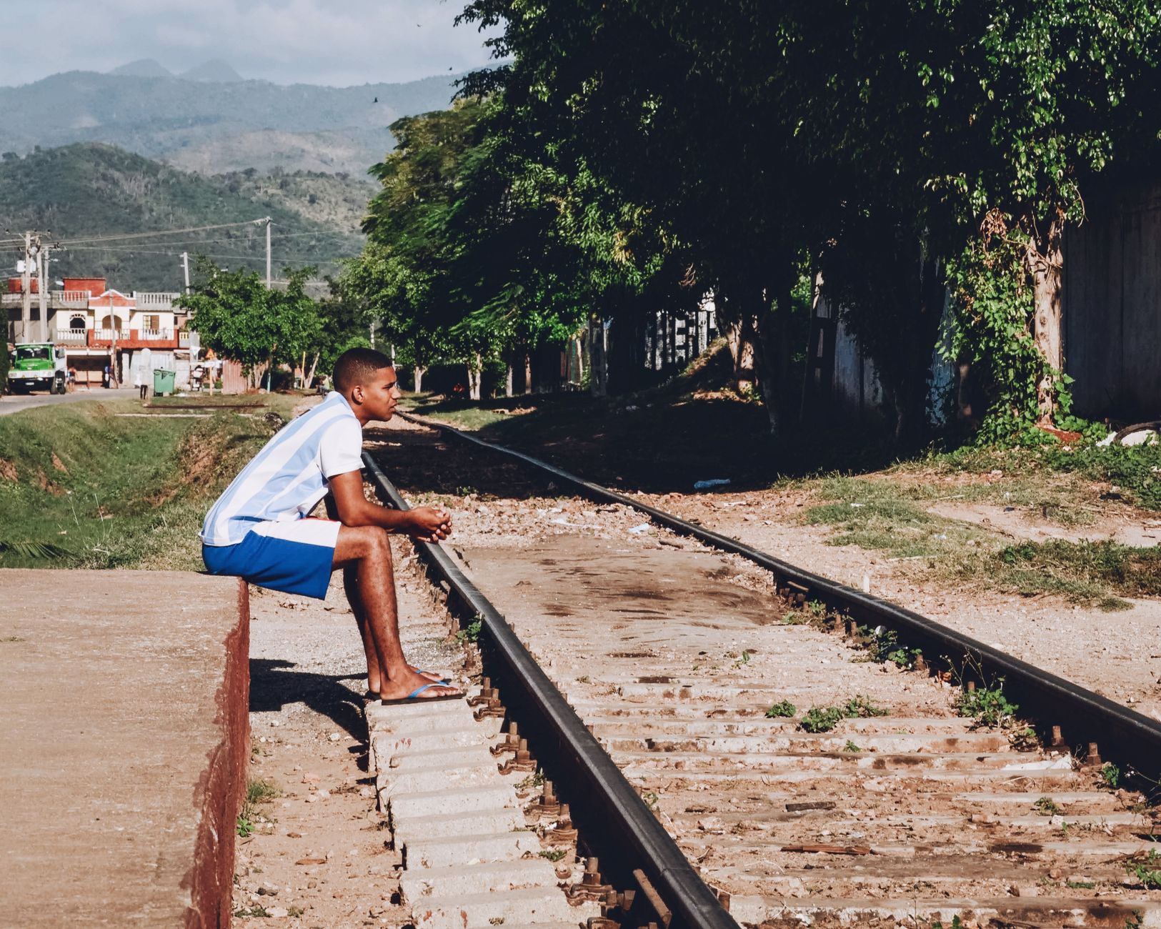A young man in casual summer attire sitting on a railway track in a sunny outdoor setting.