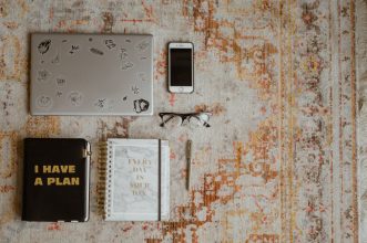 Flat lay of workspace items including laptop, phone, planners, and glasses on a patterned rug.