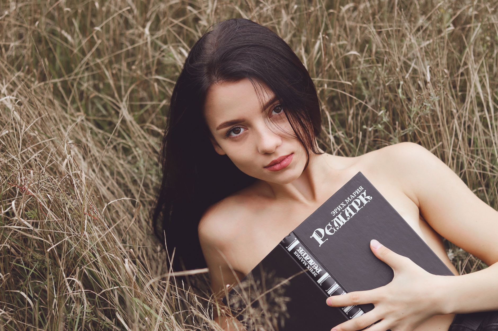 A woman lies in a field holding a book, surrounded by long grass, creating a serene and thoughtful atmosphere.