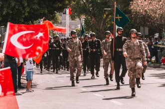 Soldiers marching in a Turkish military parade, waving national flags in a tree-lined urban setting.