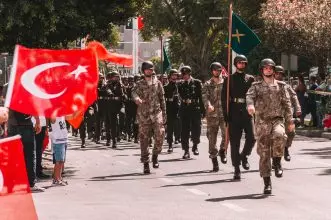 Soldiers marching in a Turkish military parade, waving national flags in a tree-lined urban setting.