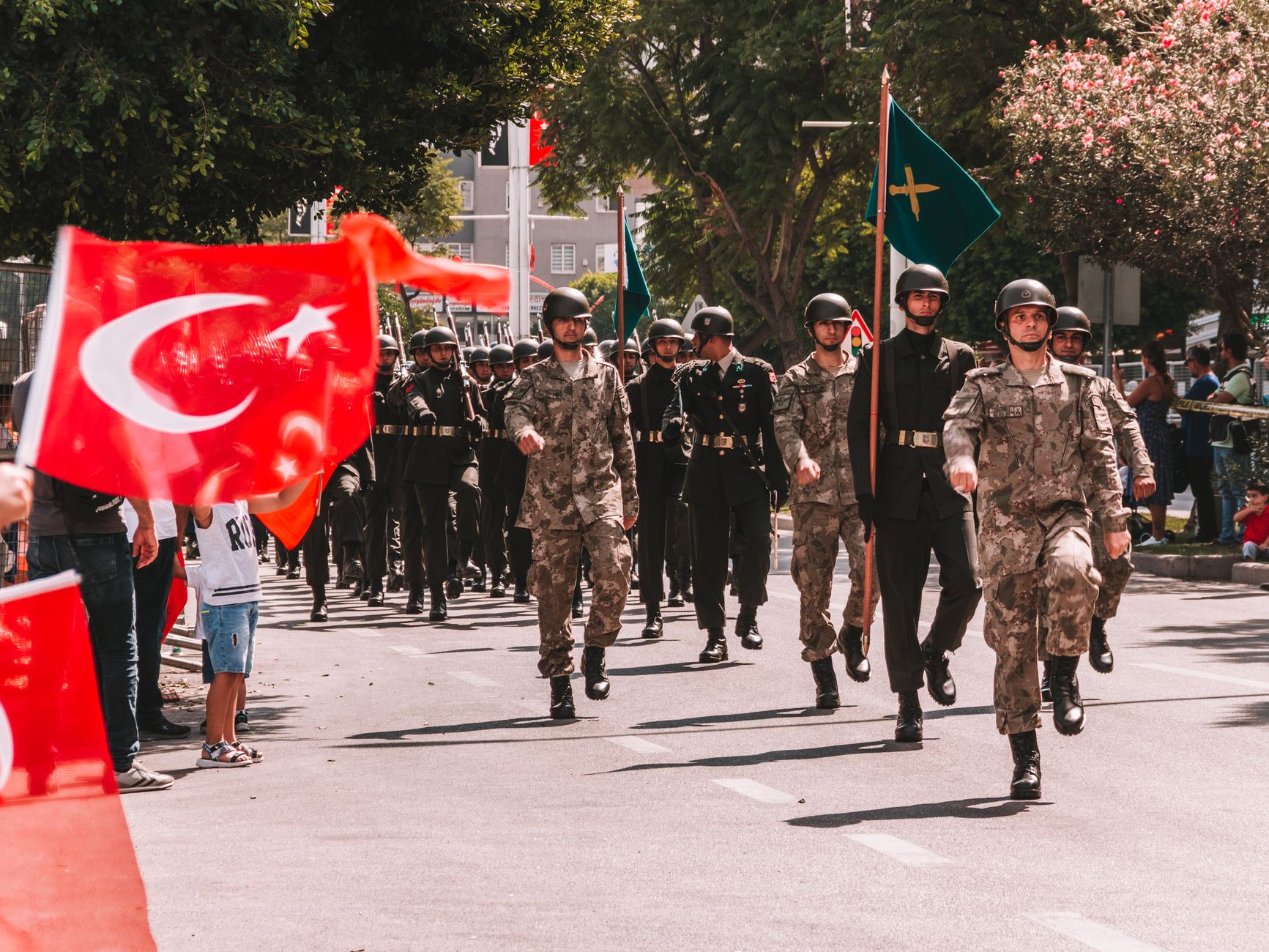 Soldiers marching in a Turkish military parade, waving national flags in a tree-lined urban setting.