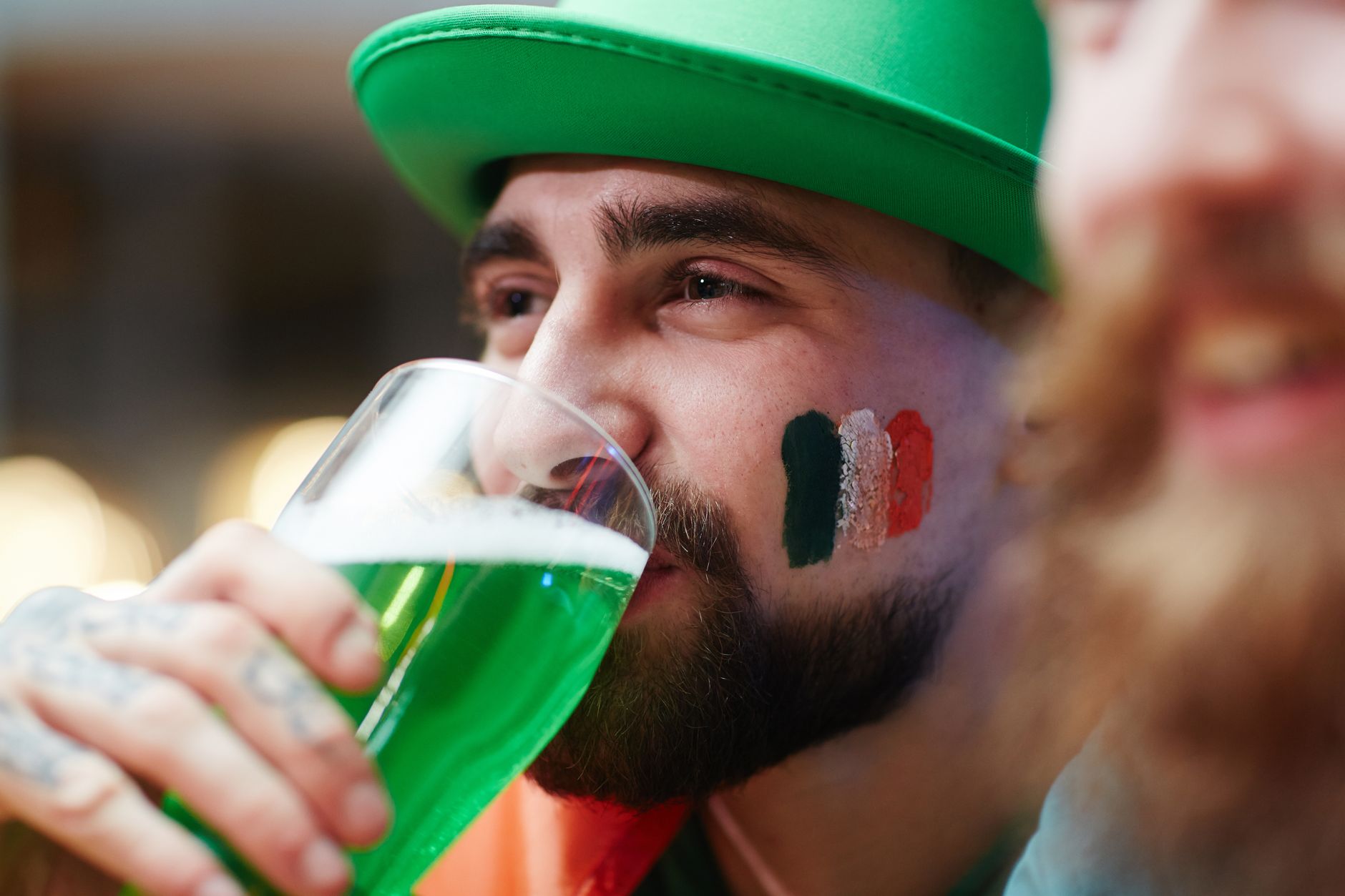 Close-up of a man enjoying green beer with an Irish flag painted on his cheek during St. Patrick's Day.